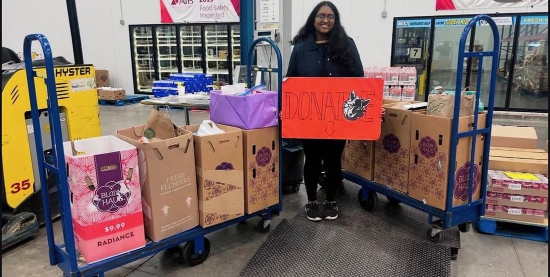 Lahari Raja standing with a hand-painted DONATE sign beside carts stacked with bags and boxes of donated food on delivery day